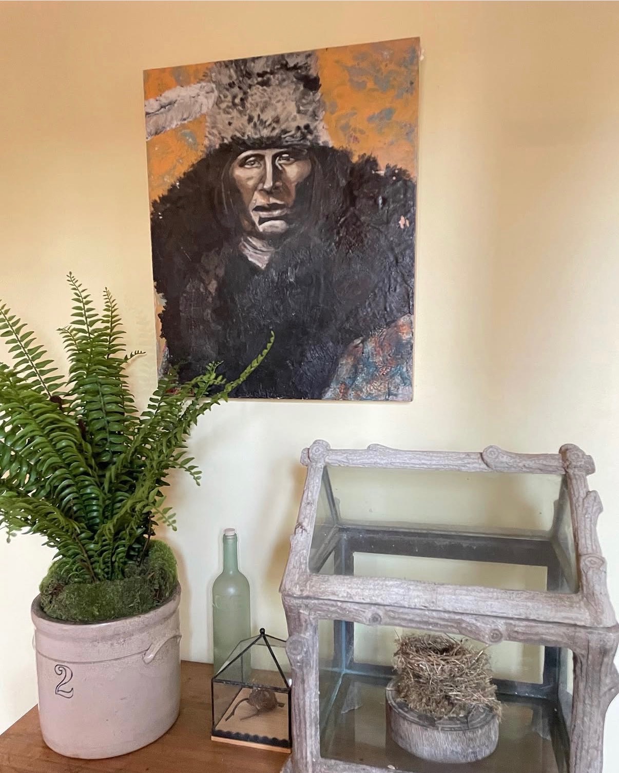 Annie Decamp portrait of an Indigenous man in a fur hat displayed on a pale yellow wall above rustic decor