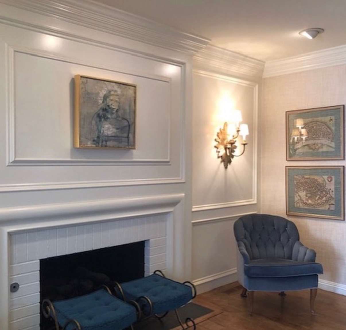 A painting above a white fireplace in an interior with blue upholstered seating and framed artwork on the adjoining wall.
